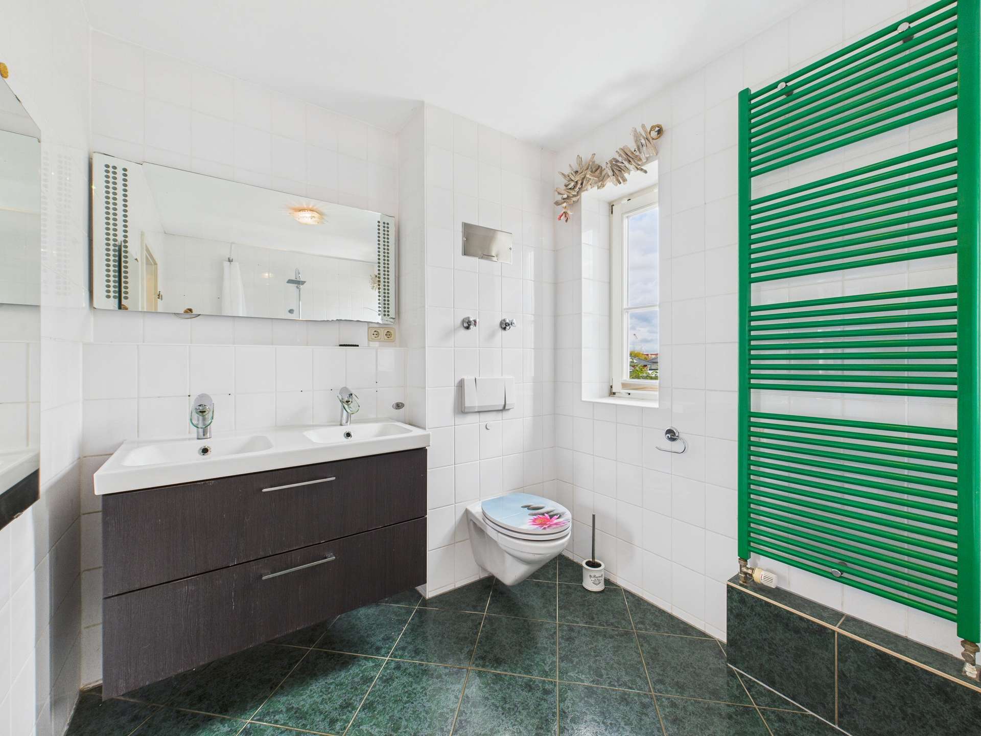 Helles Badezimmer mit Doppelwaschtisch, moderner Toilette und grüner Handtuchheizung an der Wand.