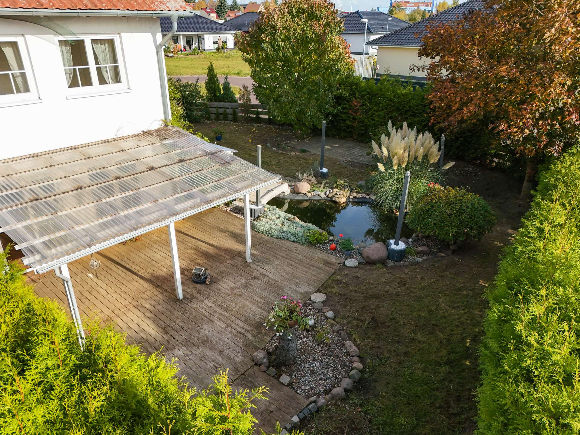 Blick auf einen Garten mit Teich, Holzterrasse und gepflegten Pflanzen, umgeben von Hecken und Bäumen.