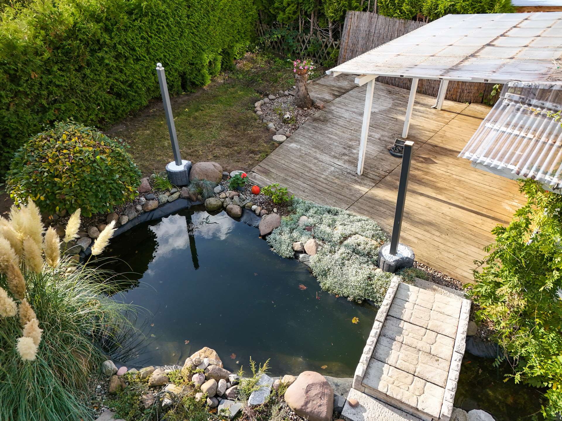 Ein gepflegter Garten mit einem Teich, umgeben von Steinen und Pflanzen, sowie einer überdachten Terrasse.