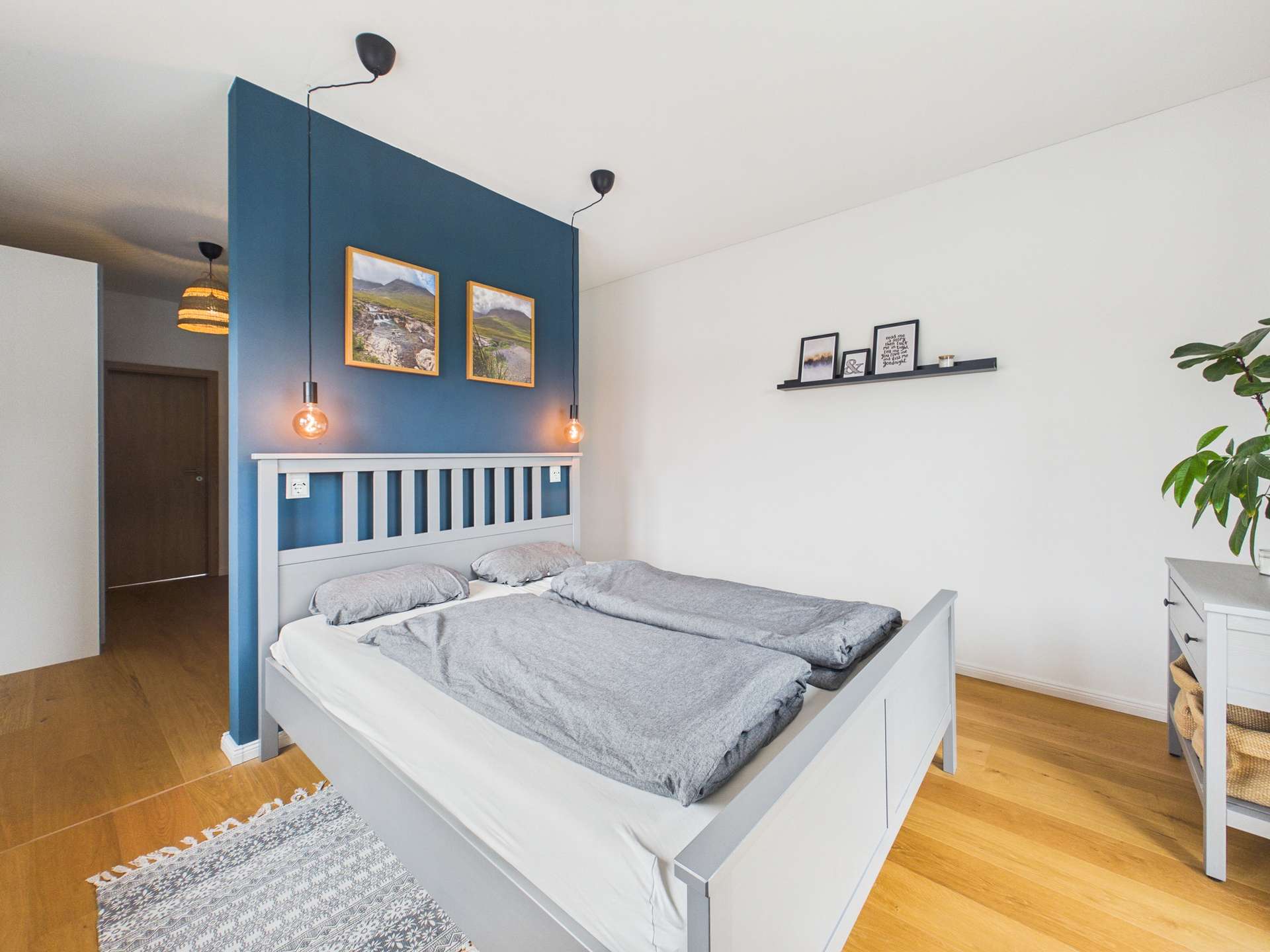 Helles Schlafzimmer mit einem Doppelbett, blauer Wand, Bildern, Regalen und einem Pflanzenarrangement.