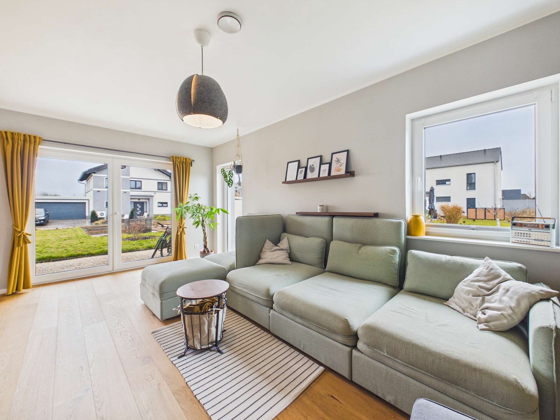 Moderner Wohnraum mit einem hellen, gemütlichen Sofa, einem runden Tisch und großen Fenstern, die auf einen Garten blicken.