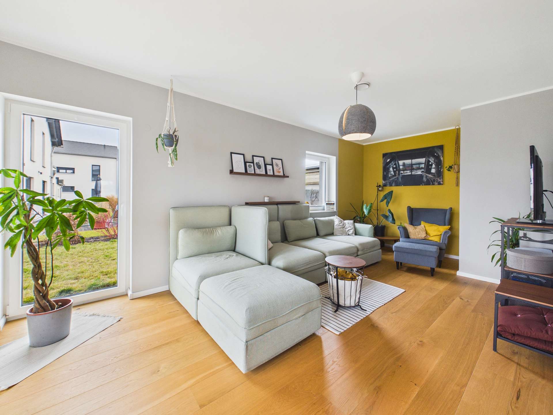 Helles Wohnzimmer mit grauem Sofa, gelber Wand, Pflanzen und moderner Einrichtung. Große Fenster mit Blick nach draußen.
