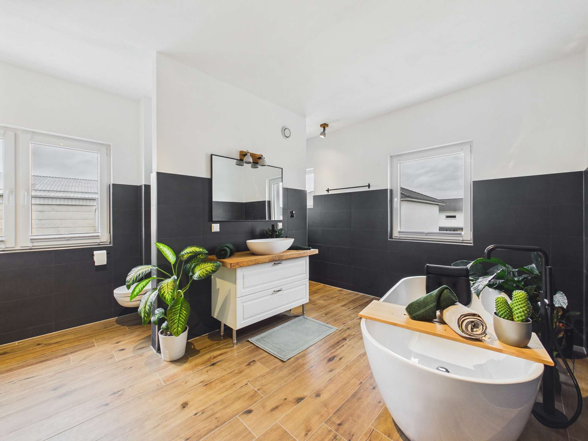 Modernes Badezimmer mit freistehender Badewanne, Holzoberfläche, Pflanzen und großen Fenstern.