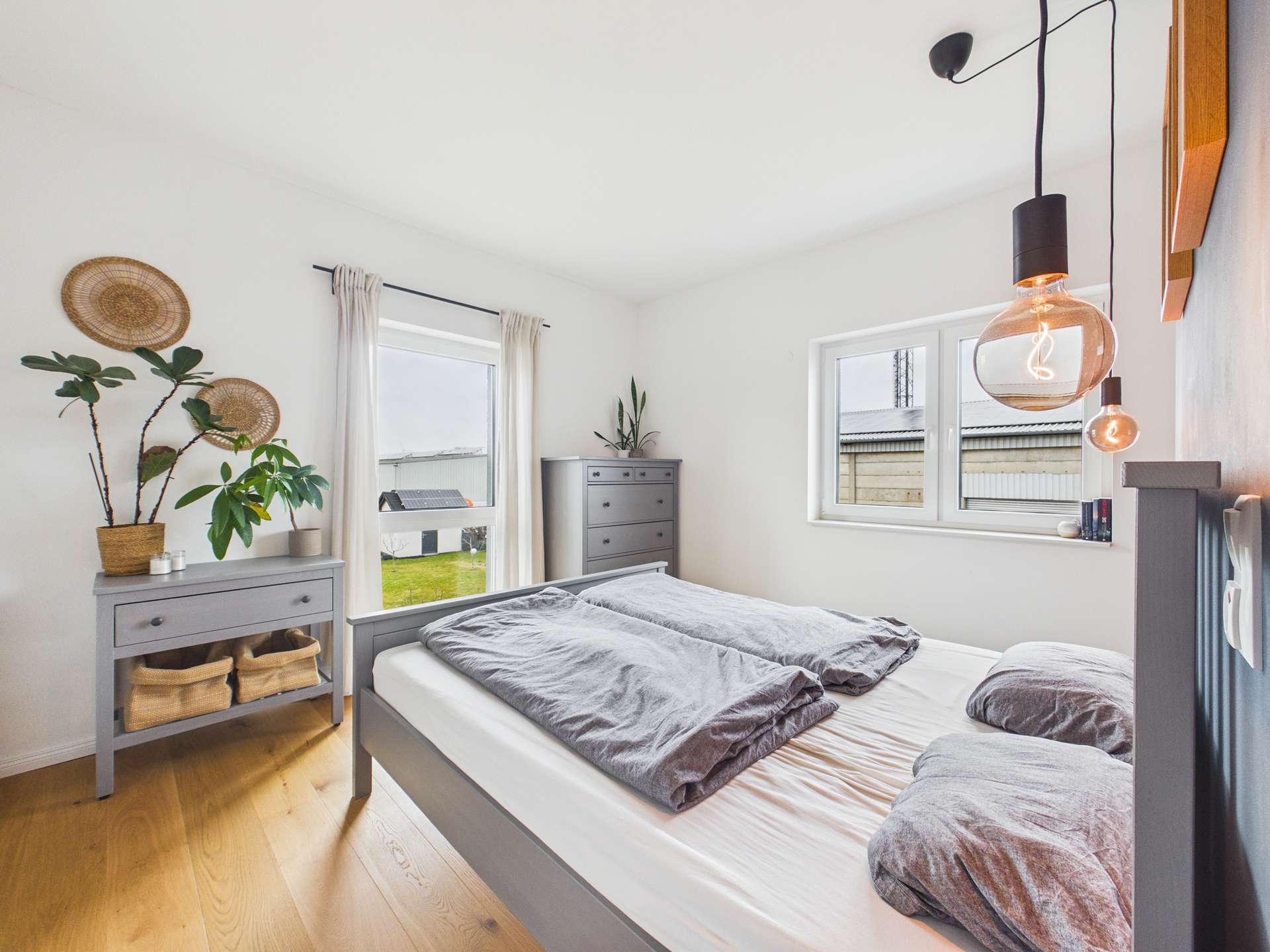 Schlafzimmer mit einem Bett, modernen Möbeln, Pflanzen und großen Fenstern, die Tageslicht hereinlassen.