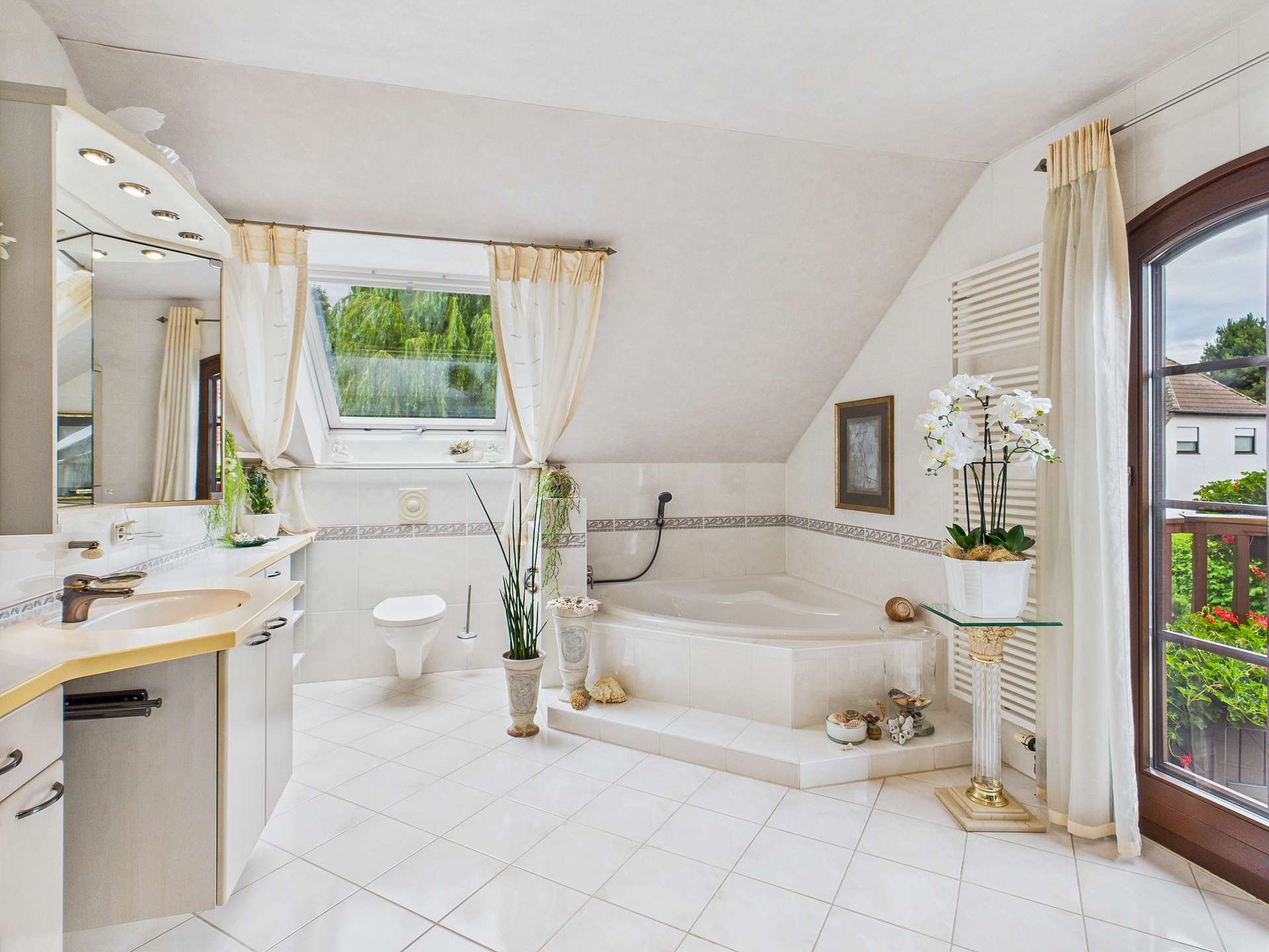 Helles Badezimmer mit Eckbadewanne, Waschbecken, Toilette und großen Fenstern, dekoriert mit Pflanzen und eleganten Accessoires.