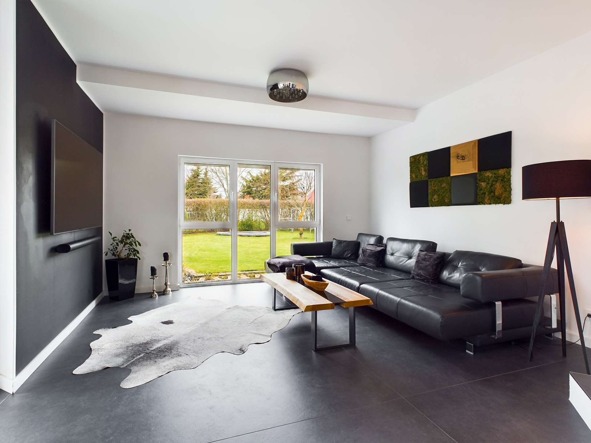 Modernes Wohnzimmer mit schwarzem Sofa, Holztisch, großen Fenstern und Blick auf den Garten.