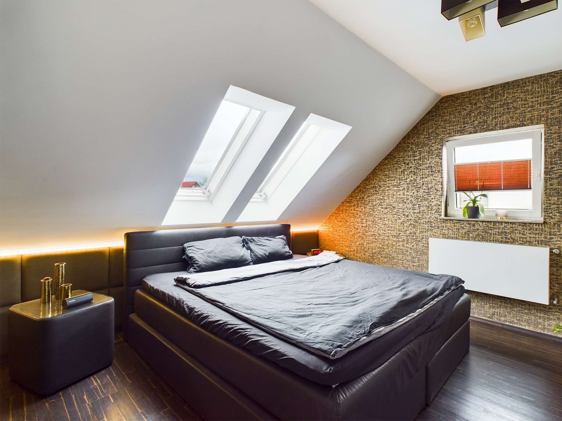 Schlafzimmer mit schrägen Fenstern, einem großen Bett mit schwarzer Bettwäsche und einer modernen Wandgestaltung.