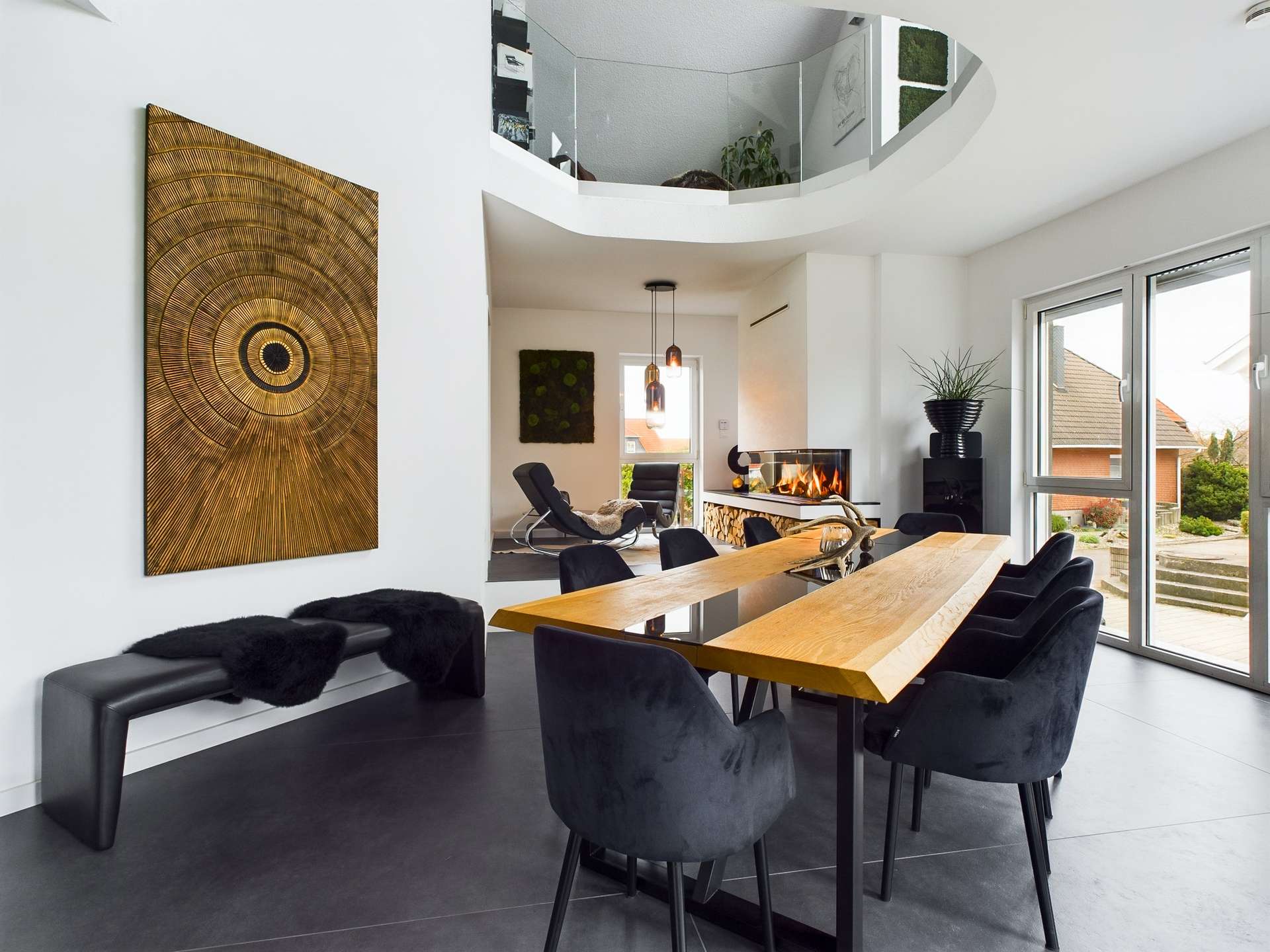 Moderne Wohnküche mit Holz-Esstisch, schwarzen Stühlen und Kunstwerk an der Wand, Kamin und großen Fenstern.