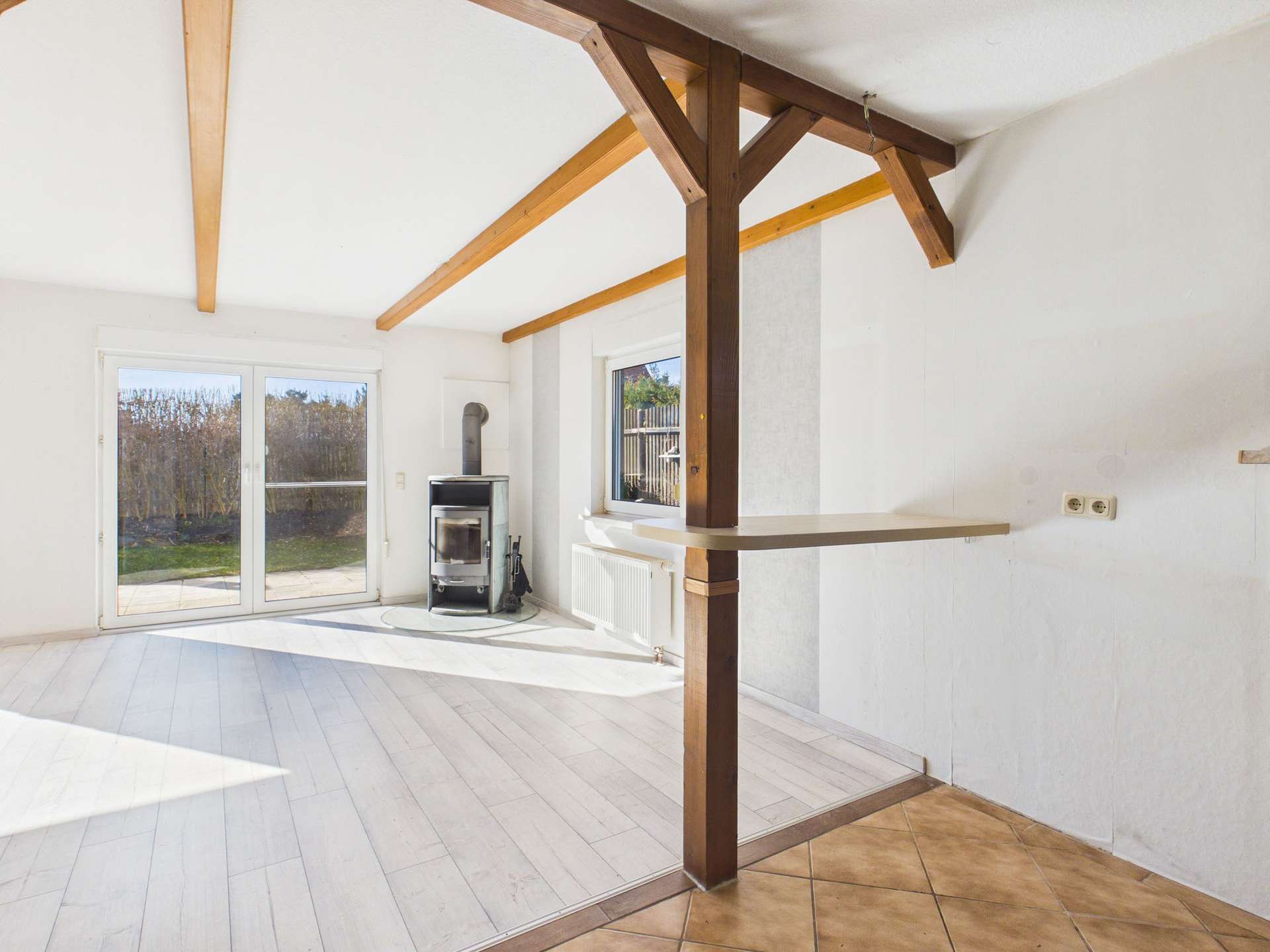 Helles, offenes Wohnzimmer mit Holzofen, großen Fenstern und Holzdecke, Blick auf den Garten.
