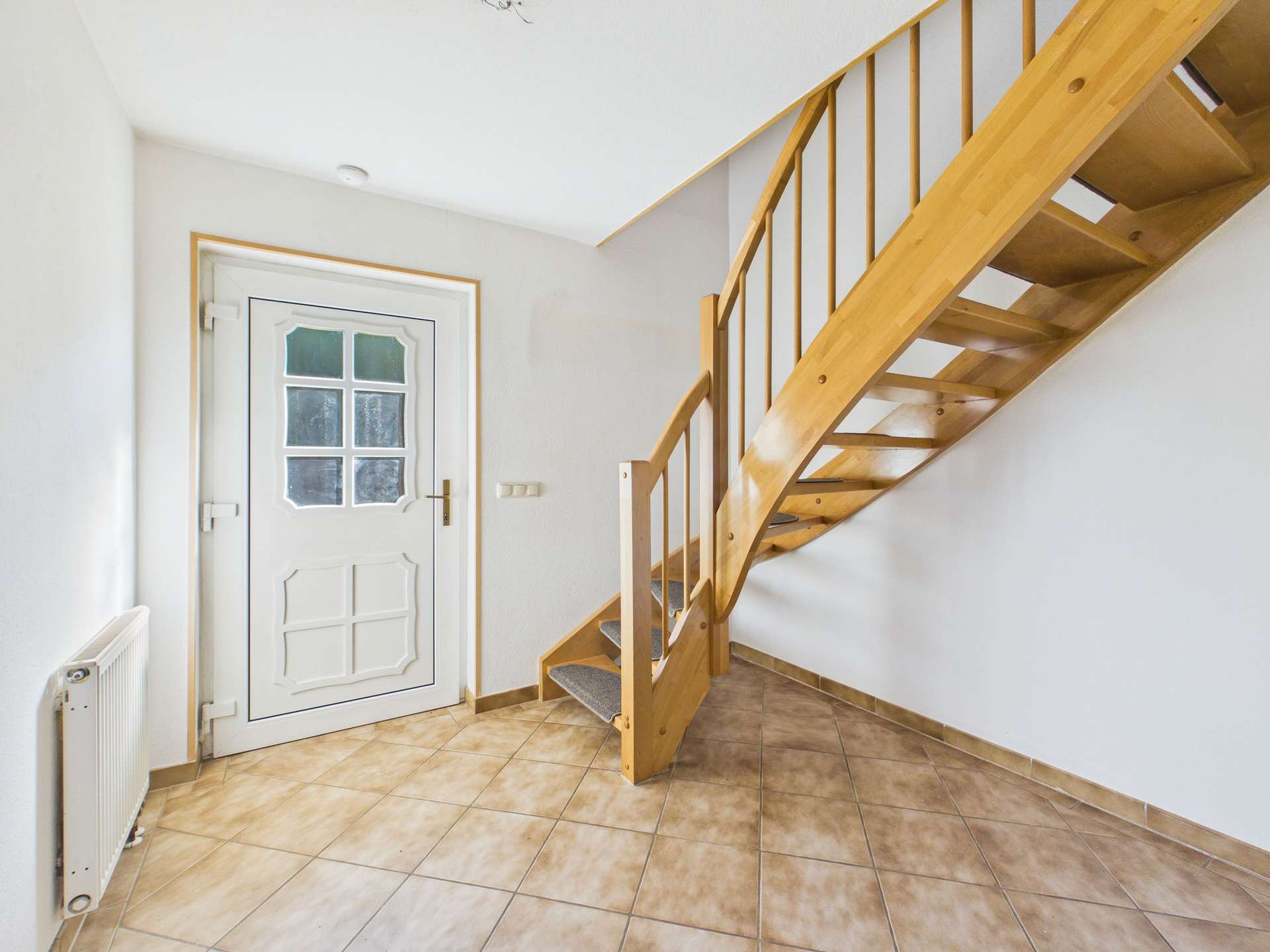 Helle Eingangshalle mit Holztreppe, Fliesenboden und weißer Tür.
