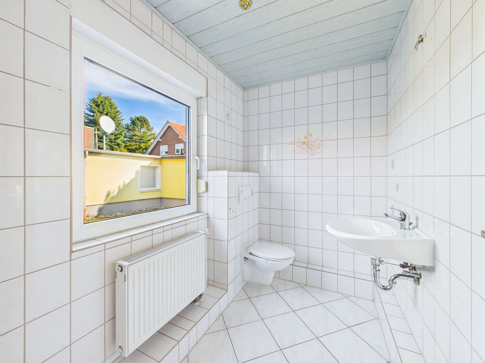 Helles Badezimmer mit weißen Fliesen, Fensterblick auf den Außenbereich, Toilette und Waschbecken.