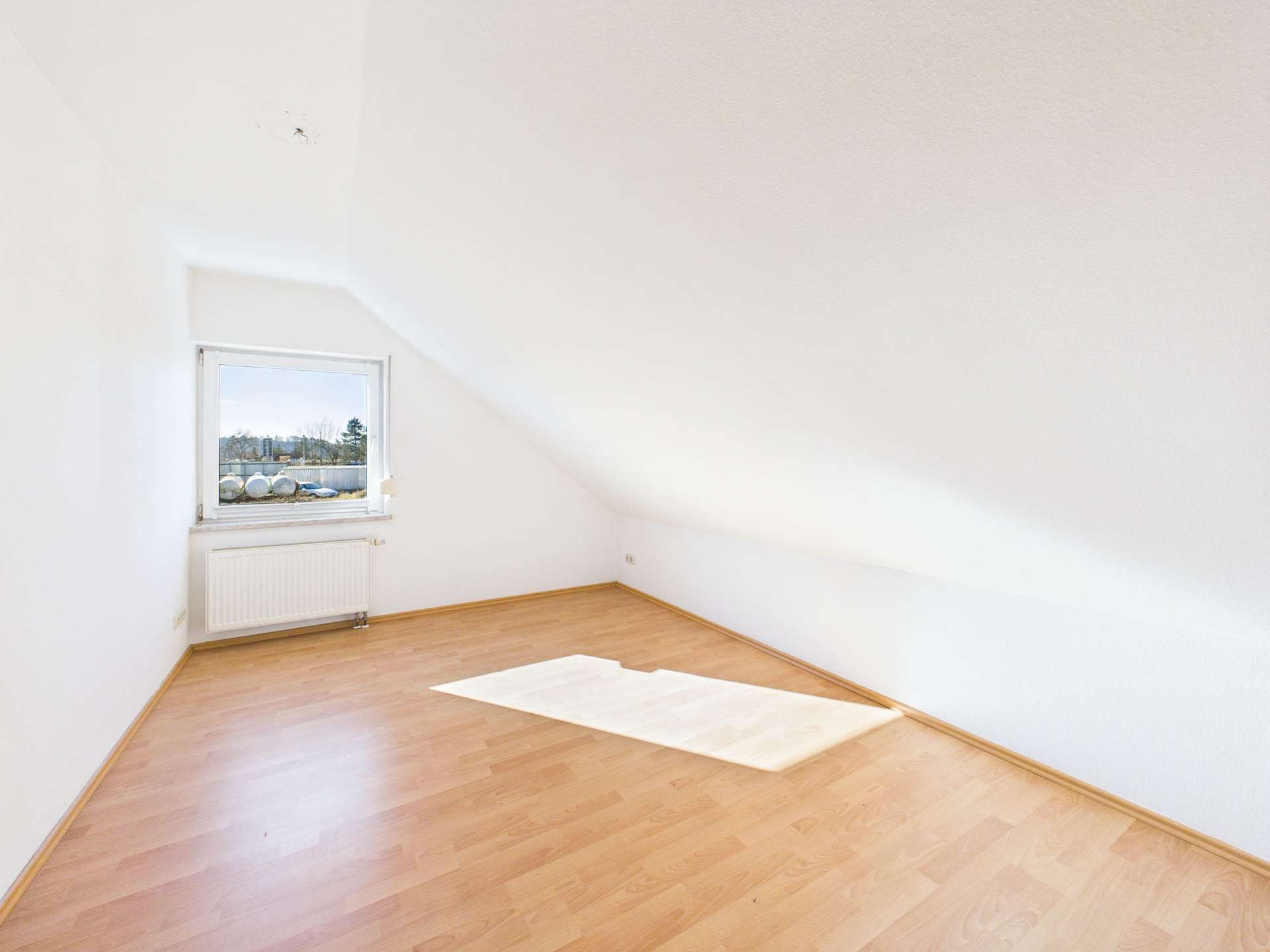 Helle, leere Dachgeschosswohnung mit Holzfußboden und Fensterblick auf eine Landschaft.