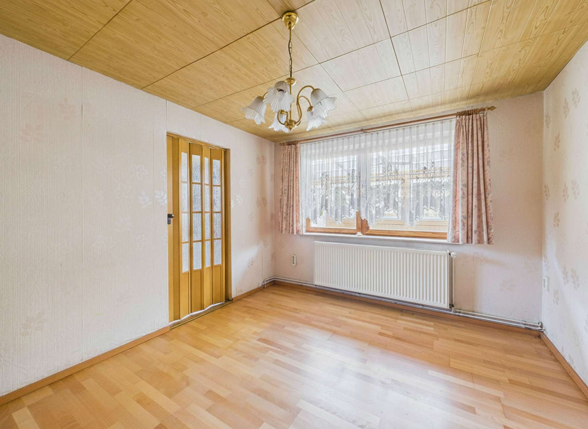 Helles Zimmer mit Holzfußboden, Fenster mit Vorhängen und einem Kronleuchter an einer Holzdecke.