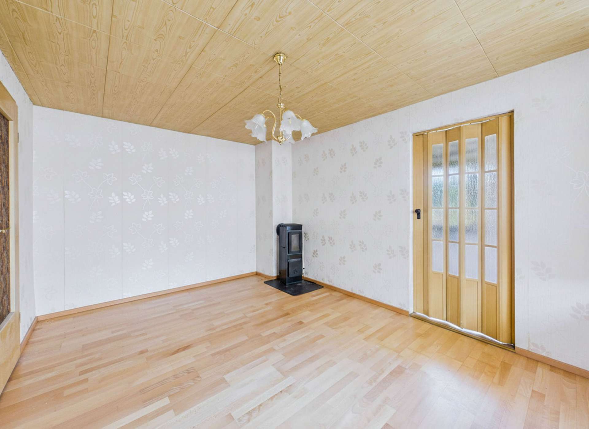 Leerer Raum mit Holzdecken, hellen Wänden, einem Ofen in der Ecke und einer Glastür mit vertikalen Lamellen.
