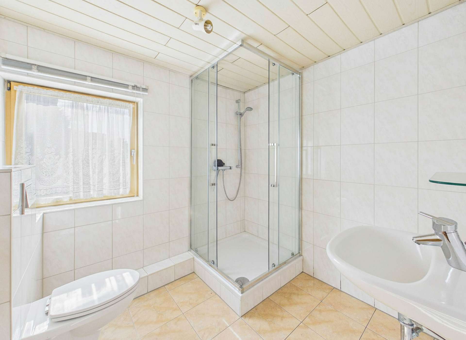 Badezimmer mit Duschkabine, Waschbecken und WC, helle Fliesen und Fenster mit Vorhang.