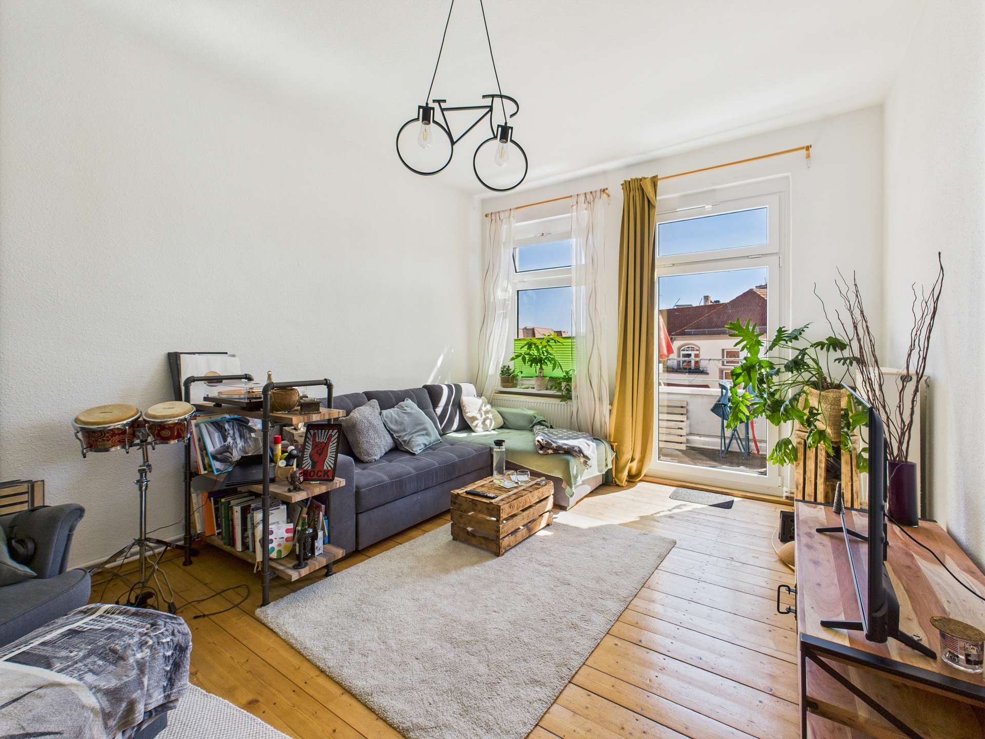 Helles Wohnzimmer mit grauem Sofa, Holztisch, Pflanzen und Fenster mit Vorhängen.