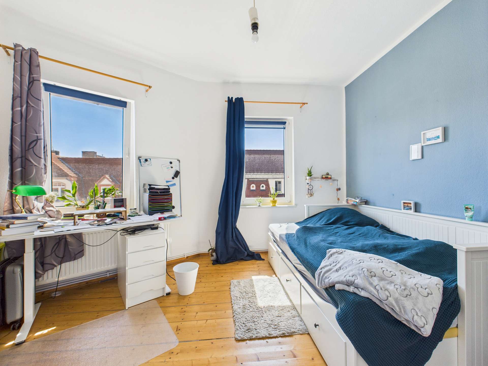 Helles Schlafzimmer mit einem Bett, Schreibtisch, Pflanzen und zwei Fenstern, die Tageslicht hereinlassen.
