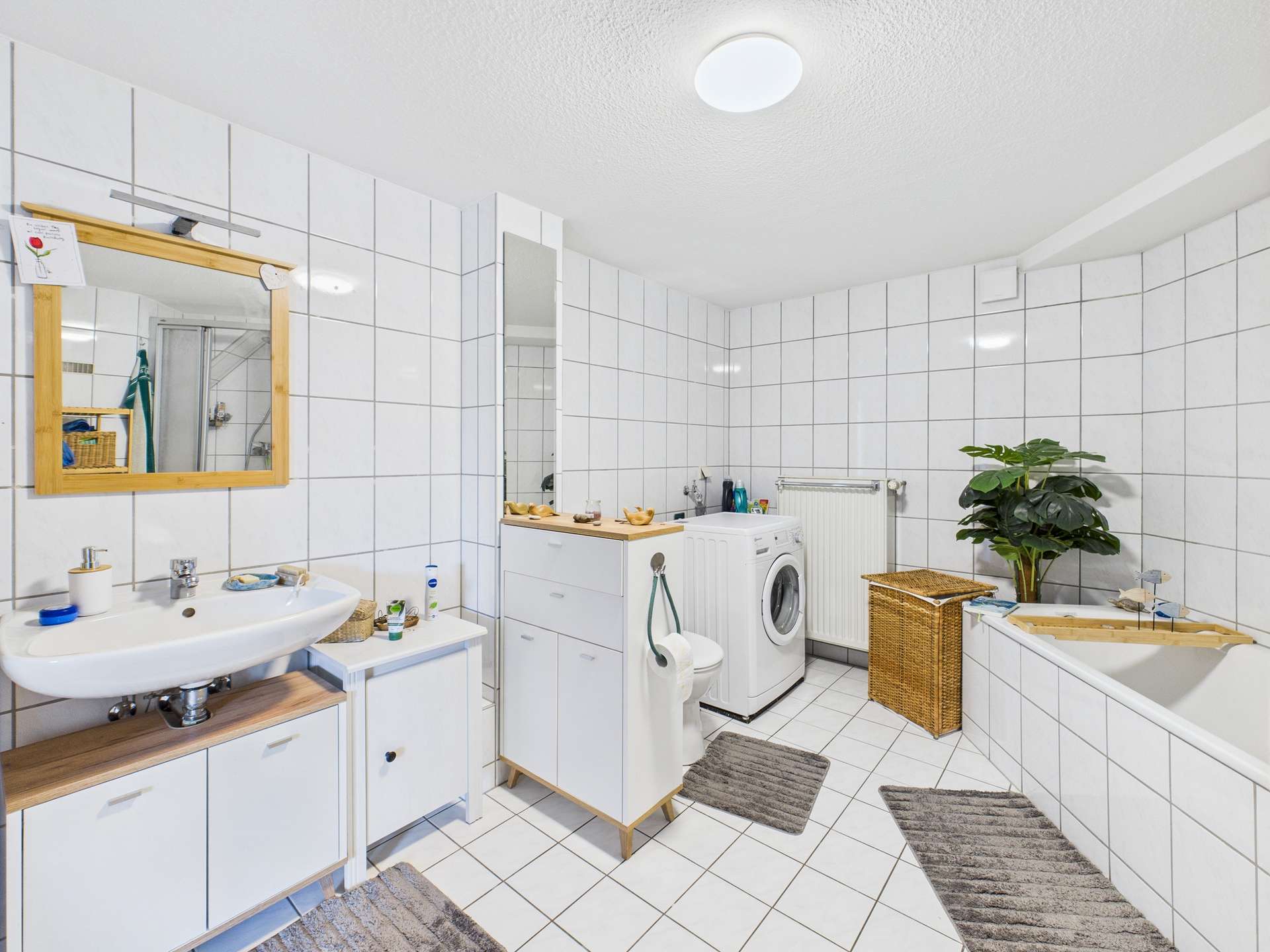Helles Badezimmer mit Waschmaschine, Waschbecken, Badewanne und Pflanzen, gefliest mit weißen Kacheln.
