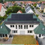Ein modernes Haus mit grünen Dachziegeln und Solarpanelen, umgeben von weiteren Wohnhäusern und einer grünen Landschaft.