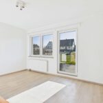 Helles, leeres Zimmer mit großen Fenstern und Blick auf einen Garten. Helle Wände und Holzboden.