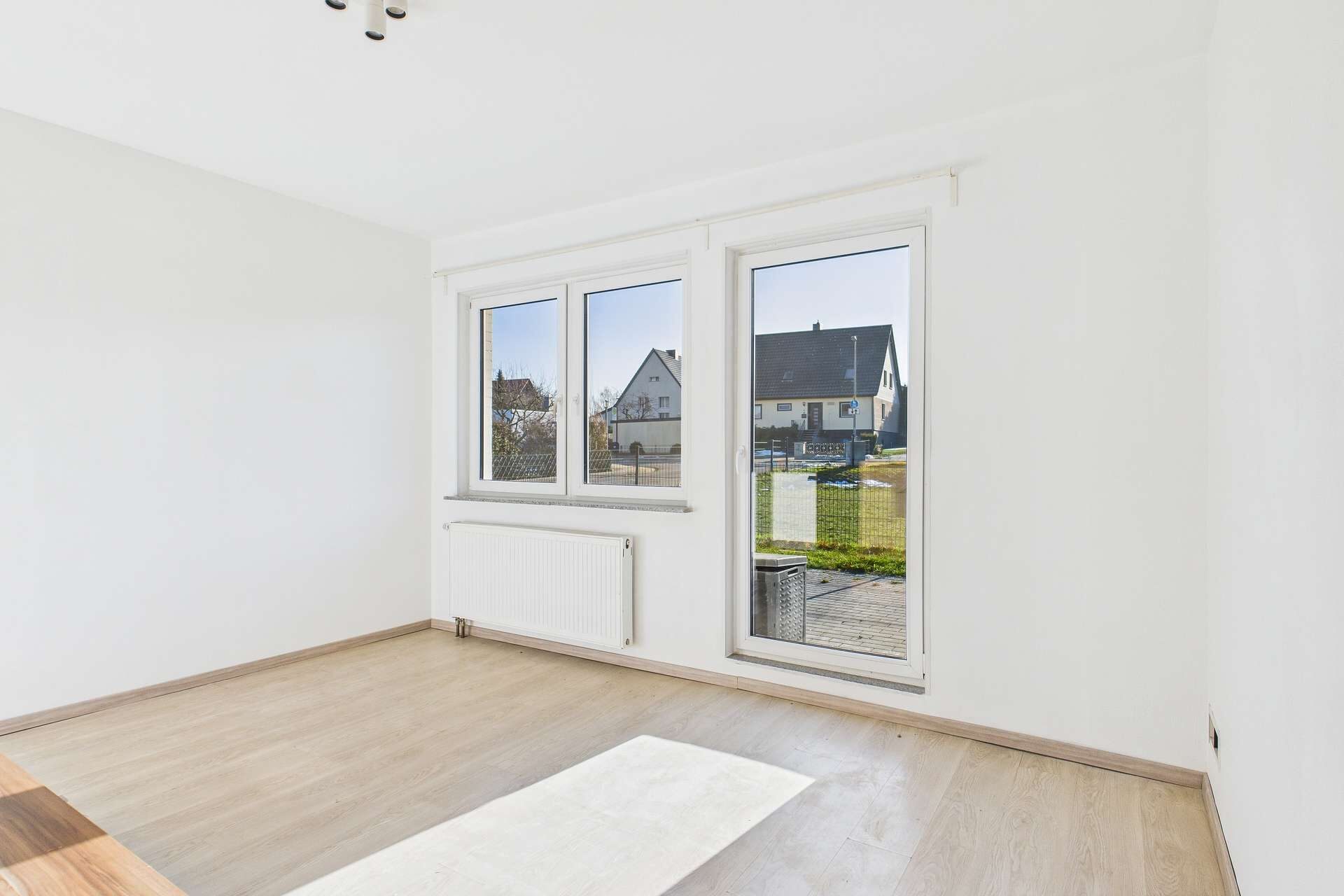 Helles, leeres Zimmer mit großen Fenstern und Blick auf einen Garten. Helle Wände und Holzboden.
