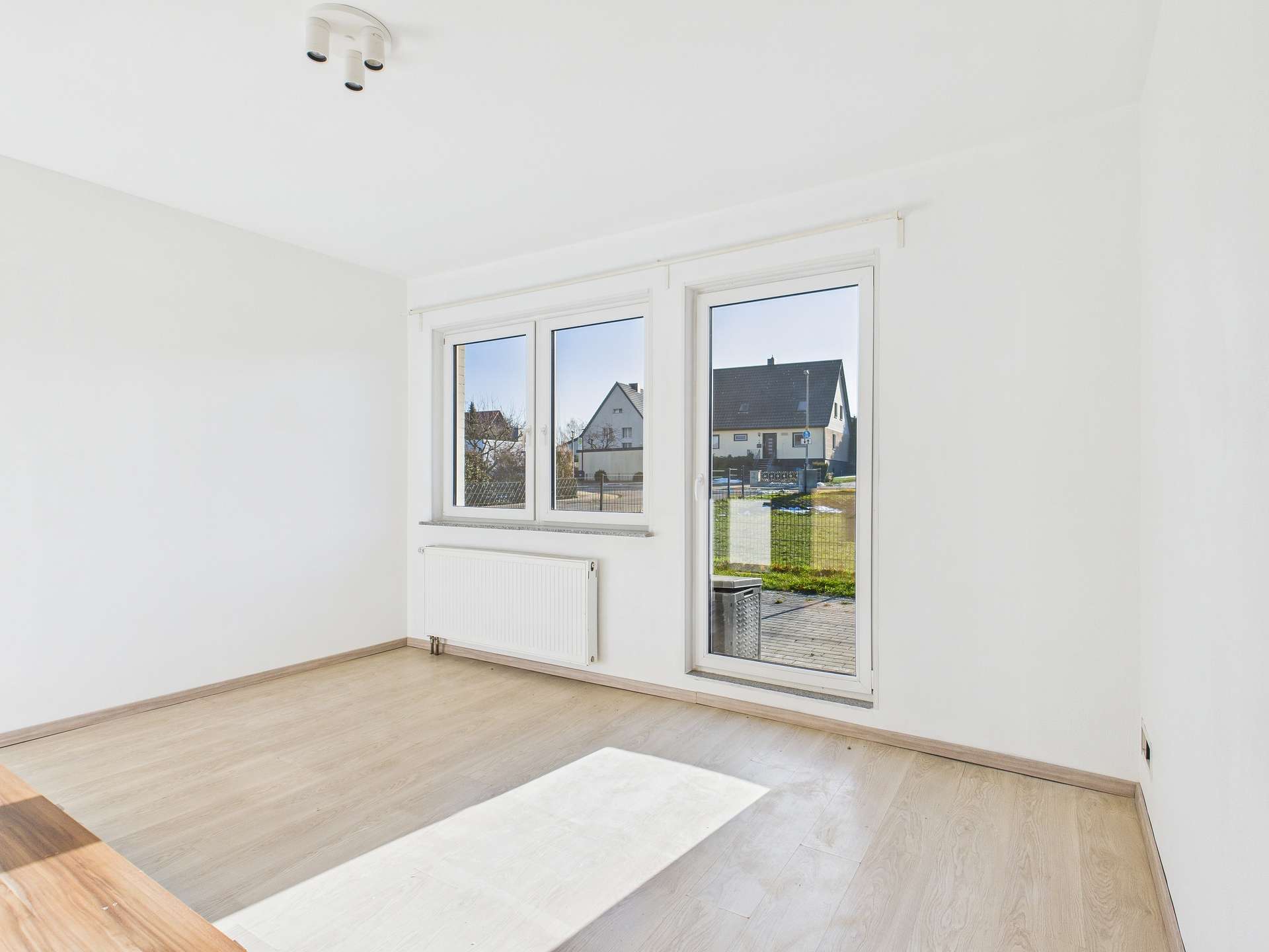 Helles, leeres Zimmer mit großen Fenstern und Blick auf einen Garten. Helle Wände und Holzboden.