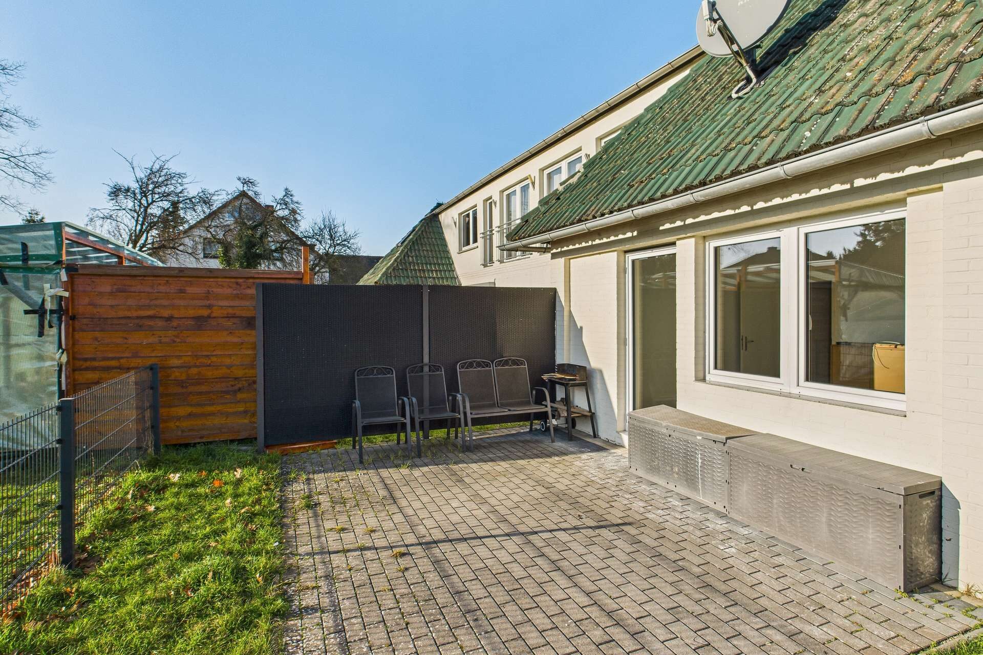 Hinterhof mit Pflastersteinen, Gartenmöbeln und einer Wand aus Sichtschutz, umgeben von einer grünen Wiese.