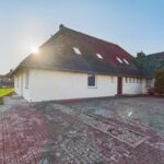 Ein einseitig geneigtes Wohnhaus mit rotem Dach, Parkplatz und Sonne im Hintergrund.