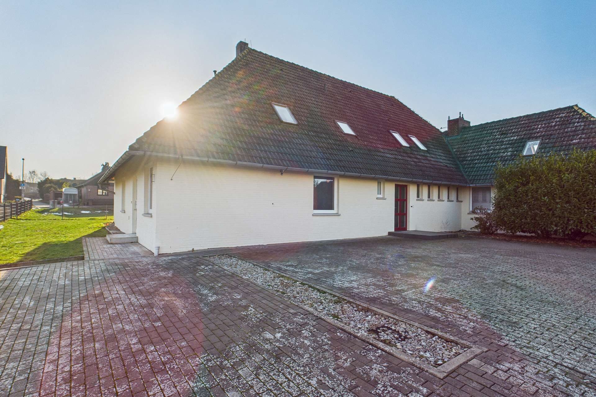 Ein einseitig geneigtes Wohnhaus mit rotem Dach, Parkplatz und Sonne im Hintergrund.