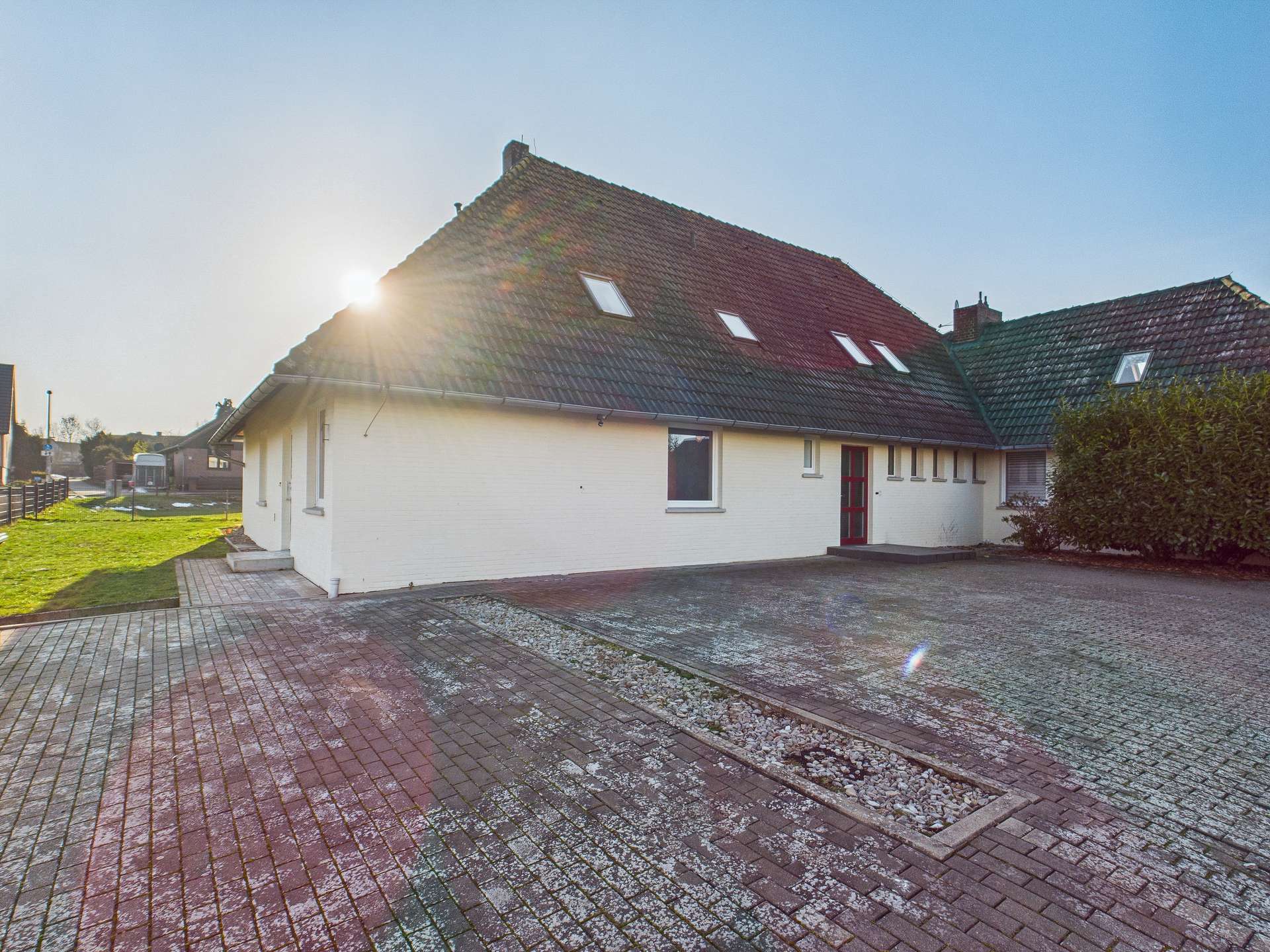 Ein einseitig geneigtes Wohnhaus mit rotem Dach, Parkplatz und Sonne im Hintergrund.
