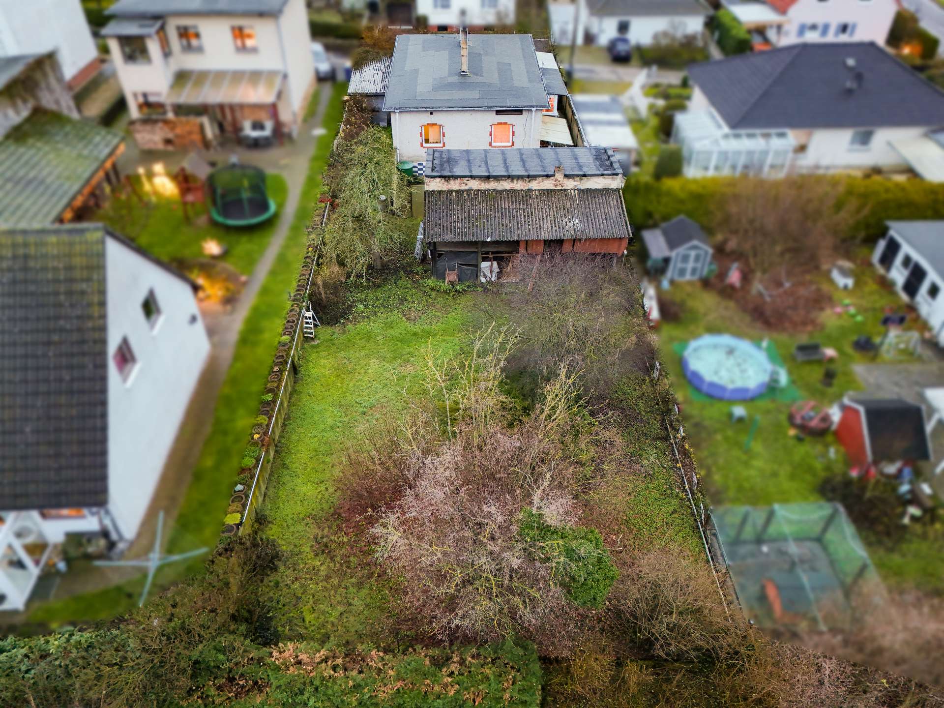 Luftaufnahme eines Gartens mit einem Haus, umgeben von Bäumen und Sträuchern, sowie einem Swimmingpool im Hintergrund.