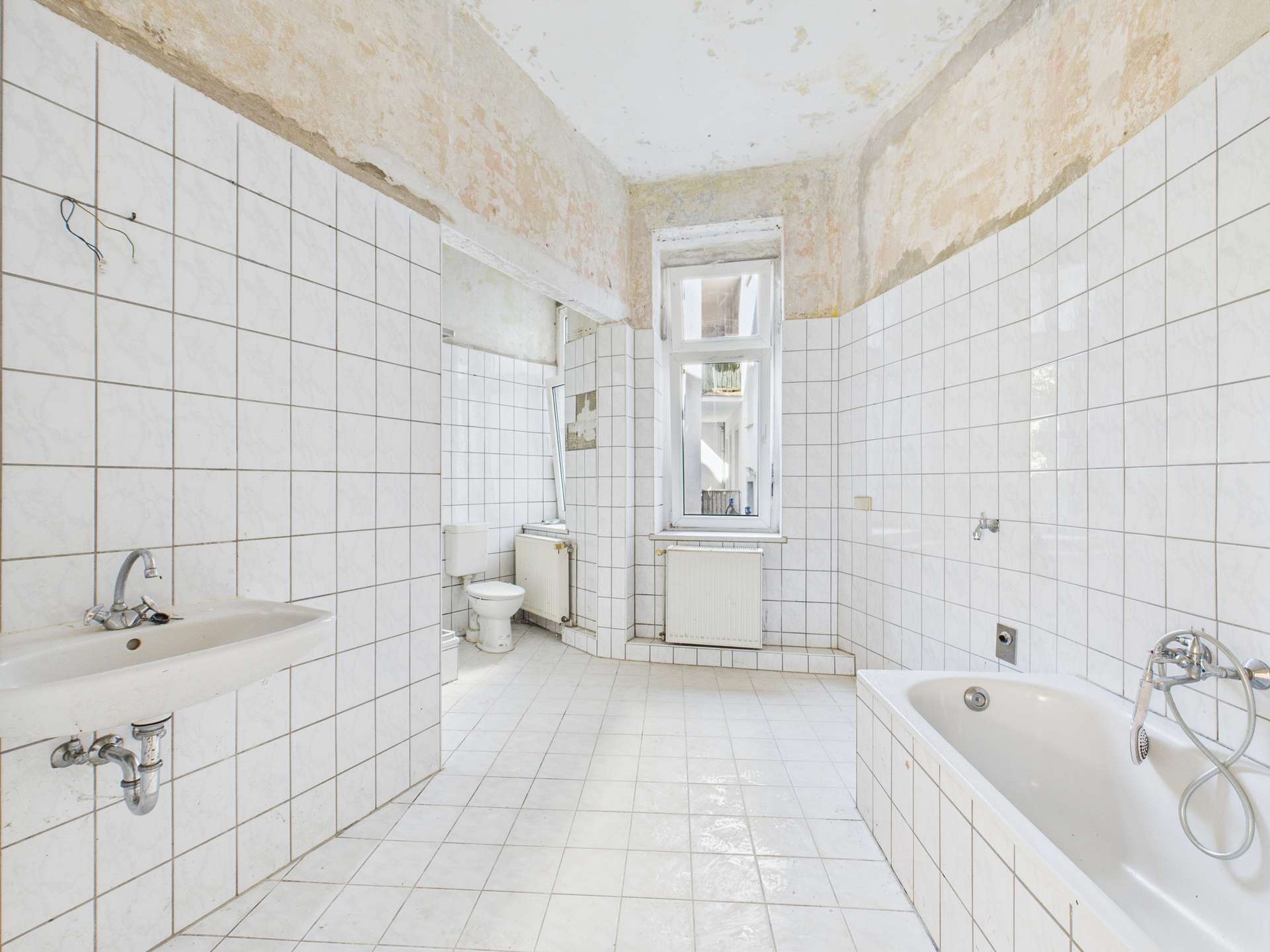 Badezimmer mit weißen Fliesen, Badewanne, Waschbecken und sichtbar abblätternder Wandfarbe.