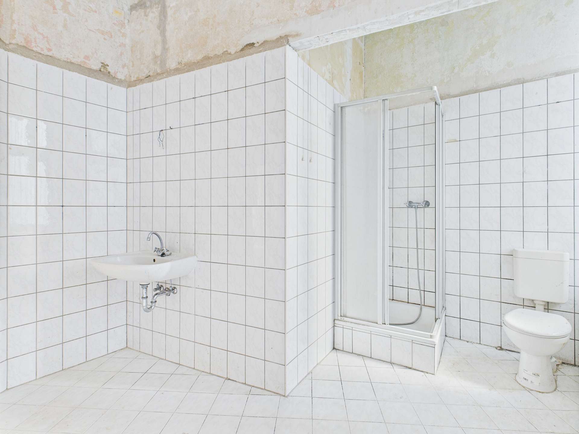 Klares, weiß gefliestes Badezimmer mit Dusche, Waschbecken und Toilette in einem renovierungsbedürftigen Zustand.