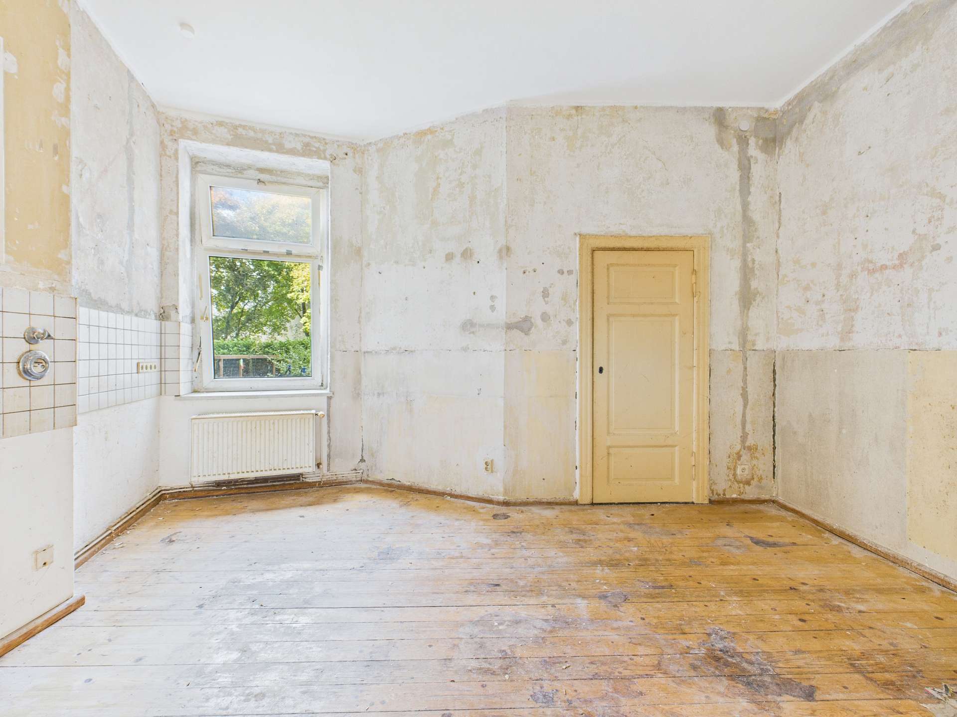 Leere, sanierungsbedürftige Wohnung mit abblätternder Wandfarbe und Holzboden, Fenster und Tür sichtbar.