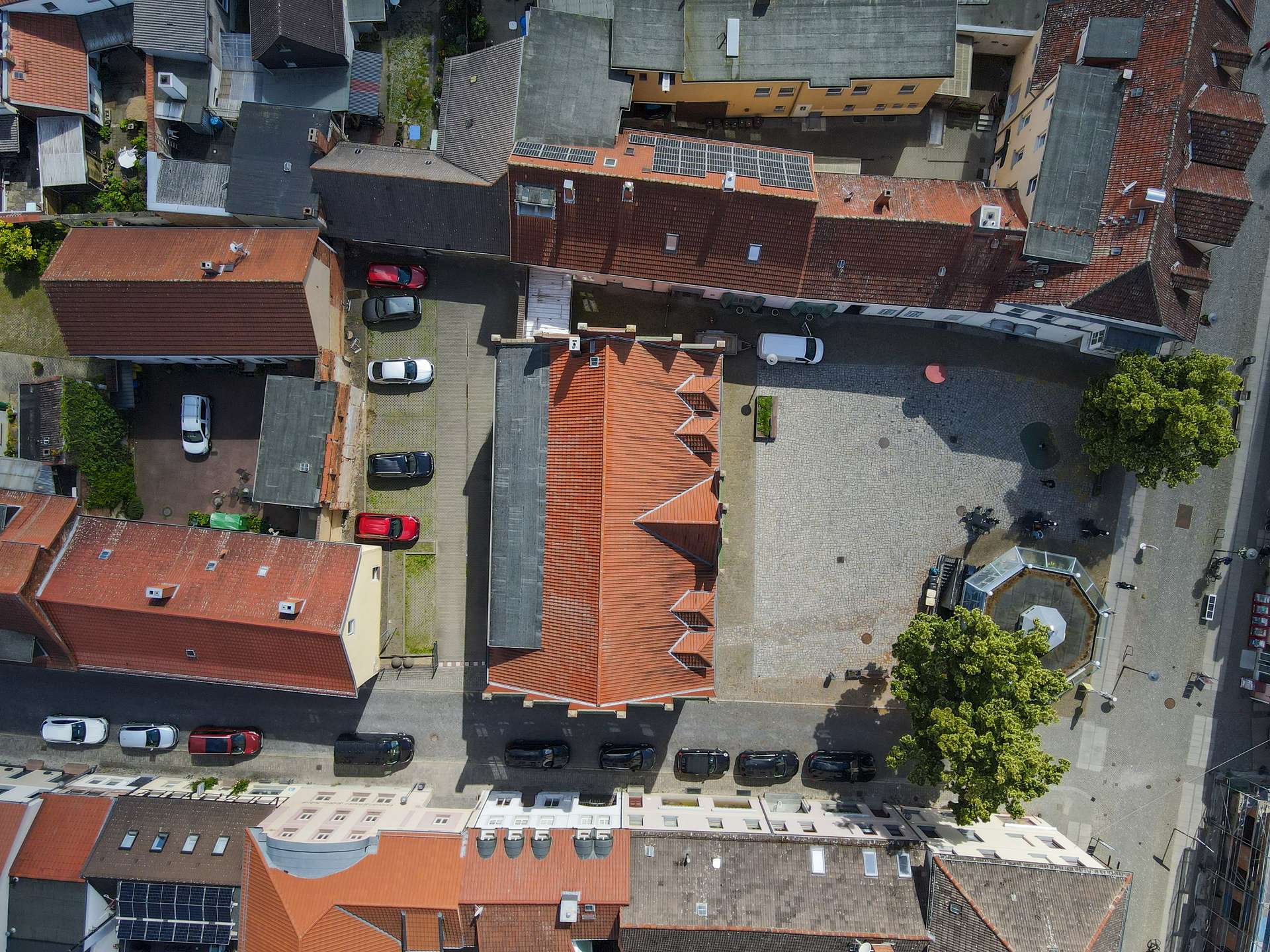 Luftaufnahme eines Stadtgebiets mit roten Dächern, Parkplätzen und einem kleinen Platz in der Mitte.
