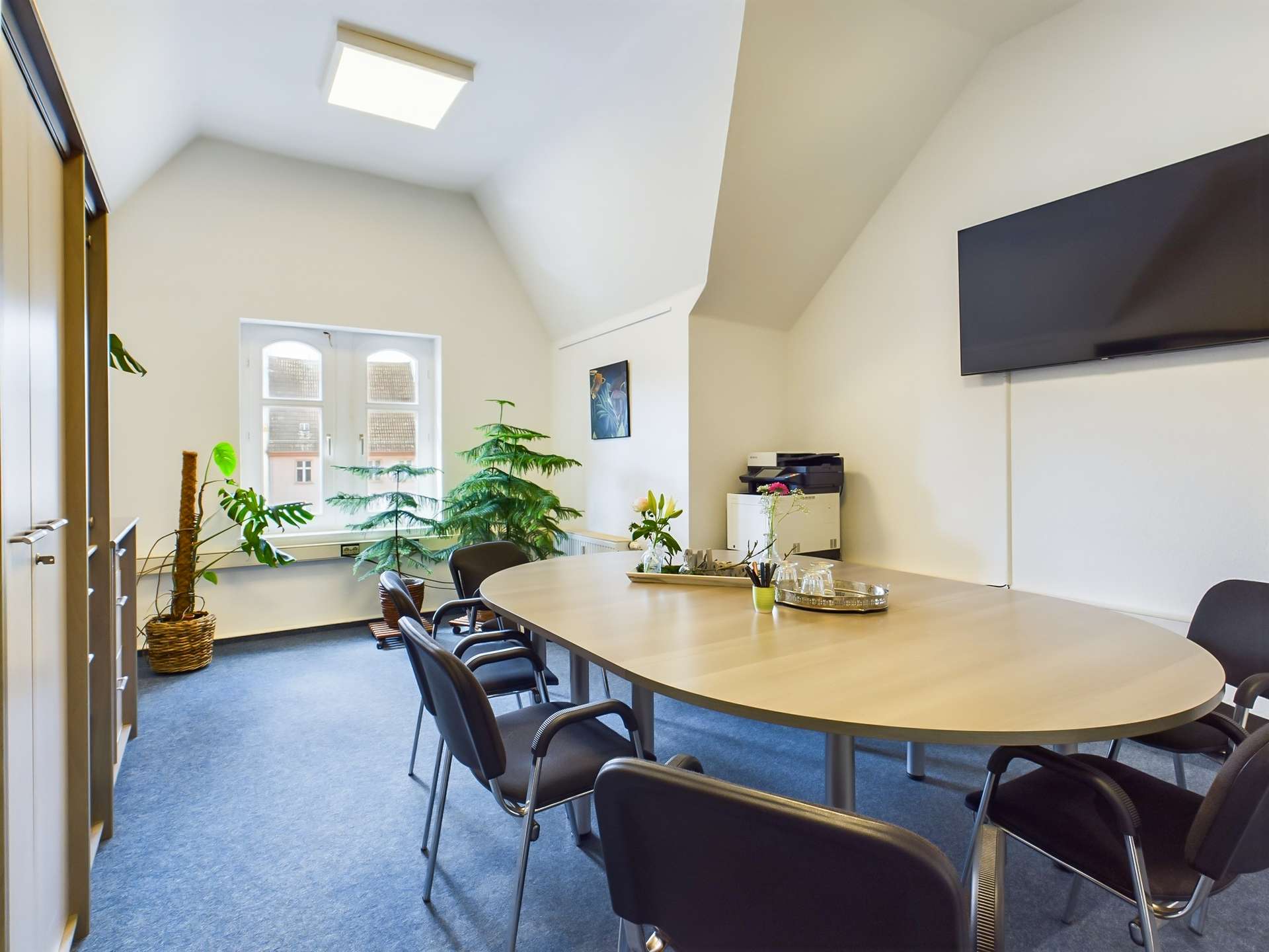 Besprechungsraum mit ovalem Tisch, schwarzen Stühlen, Pflanzen und einem Wandbild. Flachbildschirm an der Wand.