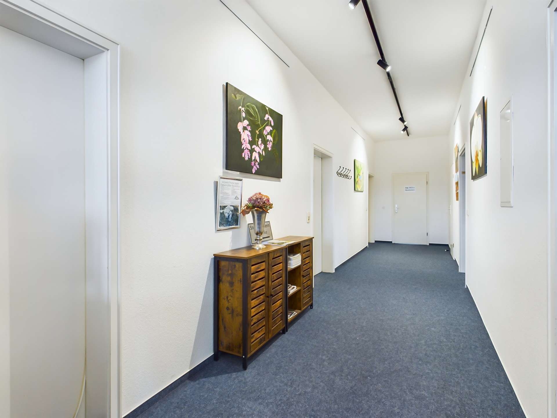 Langer, heller Flur mit blauen Teppichboden, Gemälden an der Wand und einer Holzkommode mit Blumenarrangement.