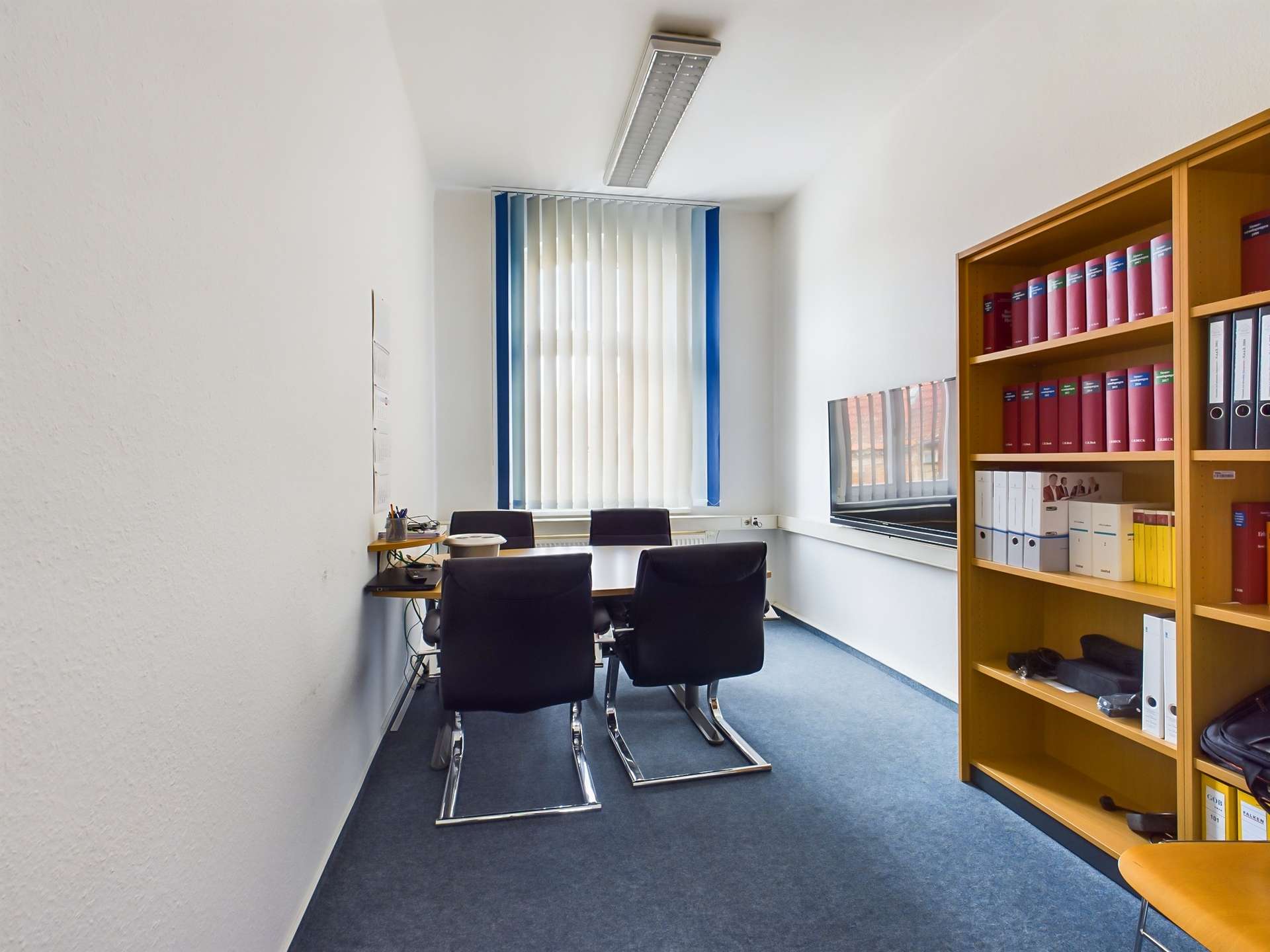 Ein Büro mit einem Konferenztisch, vier schwarzen Stühlen und einem Regal voller Aktenordner.
