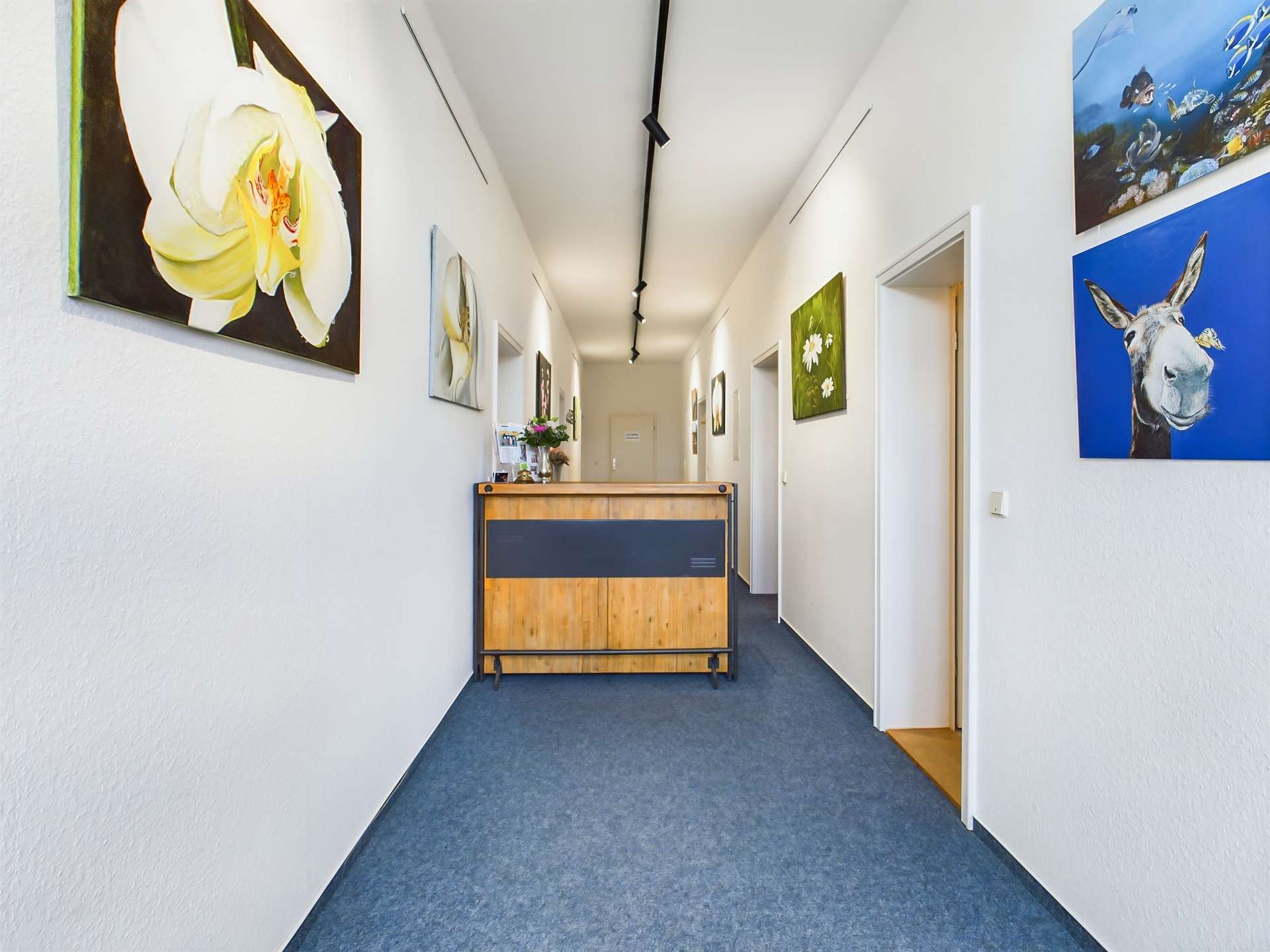 Heller Flur mit Holzempfangstresen und Bildern an den Wänden, blauer Teppichboden, moderne Atmosphäre.