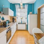 Moderne KĂŒche mit blauen WĂ€nden, Holzregalen, weiĂen SchrĂ€nken und einer Essgelegenheit am Fenster.