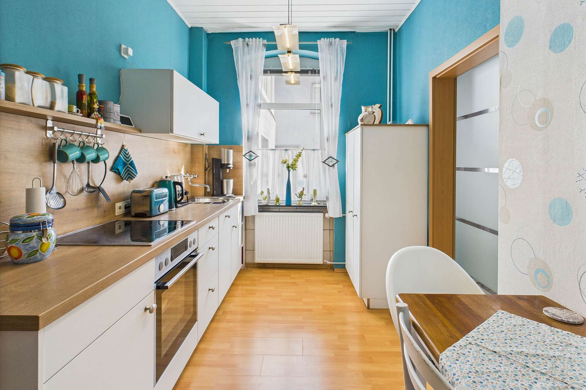 Moderne Küche in Blau und Holz mit Fenstern, hellen Vorhängen und einem kleinen Essbereich.
