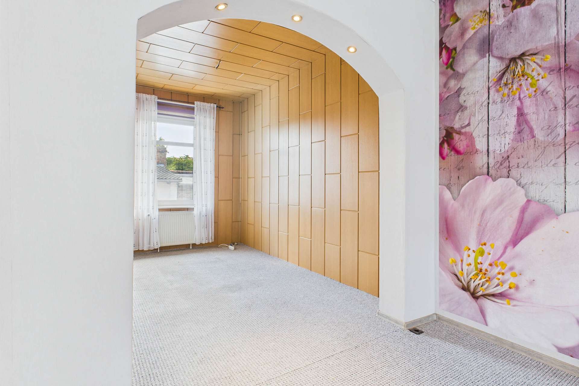 Heller Raum mit HolzvertÀfelung, Fenster mit VorhÀngen und einer Wanddekoration aus Blumenmotiven.