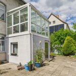 Ein heller Wintergarten an einem WohngebĂ€ude mit Terrasse und bepflanzten Töpfen, umgeben von grĂŒner Vegetation.