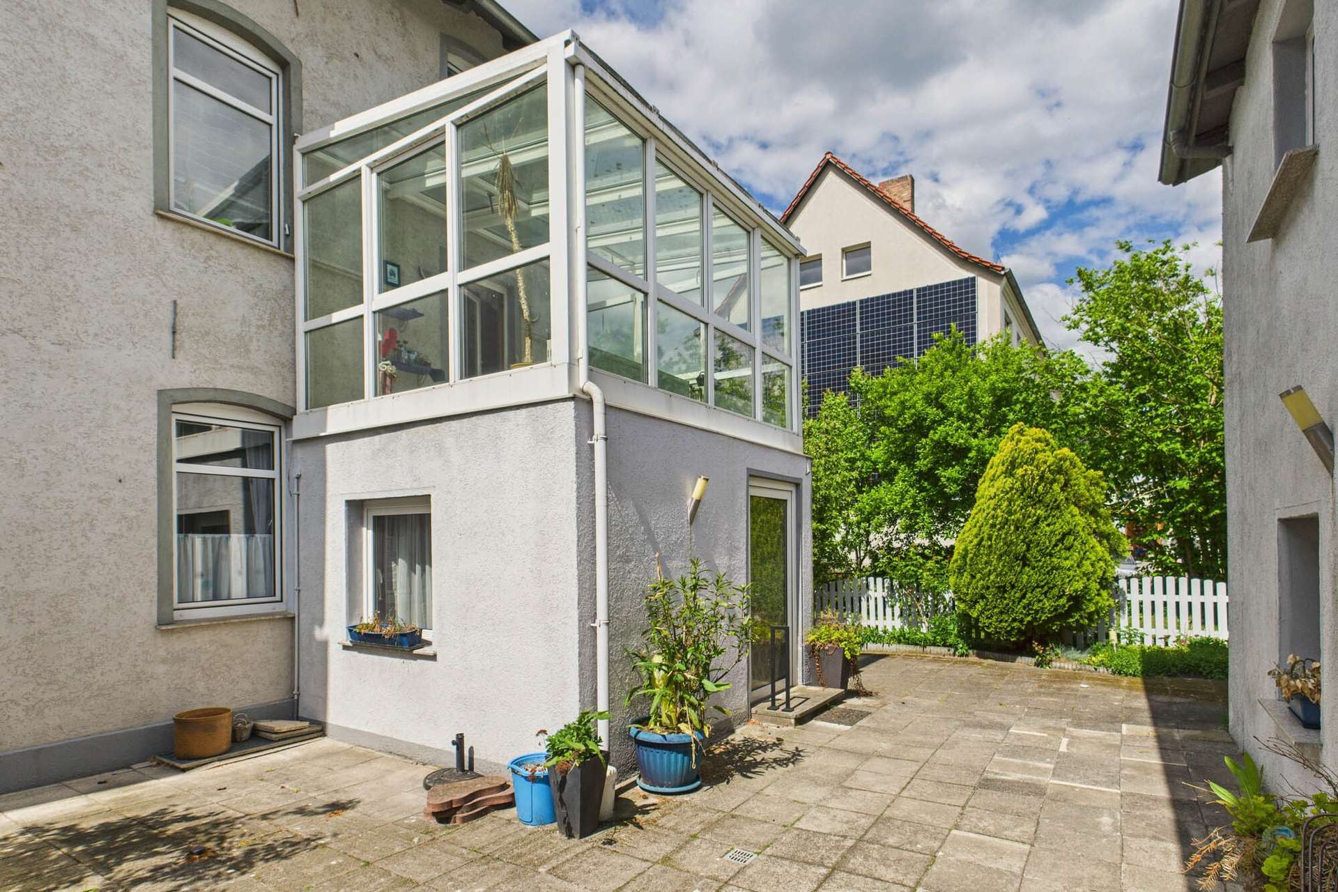 Ein heller Wintergarten an einem WohngebĂ€ude mit Terrasse und bepflanzten Töpfen, umgeben von grĂŒner Vegetation.