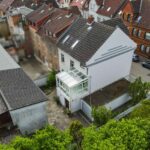 Luftaufnahme eines modernen Hauses mit Wintergarten, umgeben von traditionellen GebĂ€uden und grĂŒner Vegetation.
