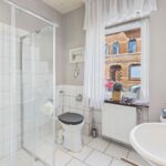 Modernes Badezimmer mit Dusche, WC, Waschbecken und Fensterblick auf eine StraĂe. Helle Fliesen und Fensterdekoration.