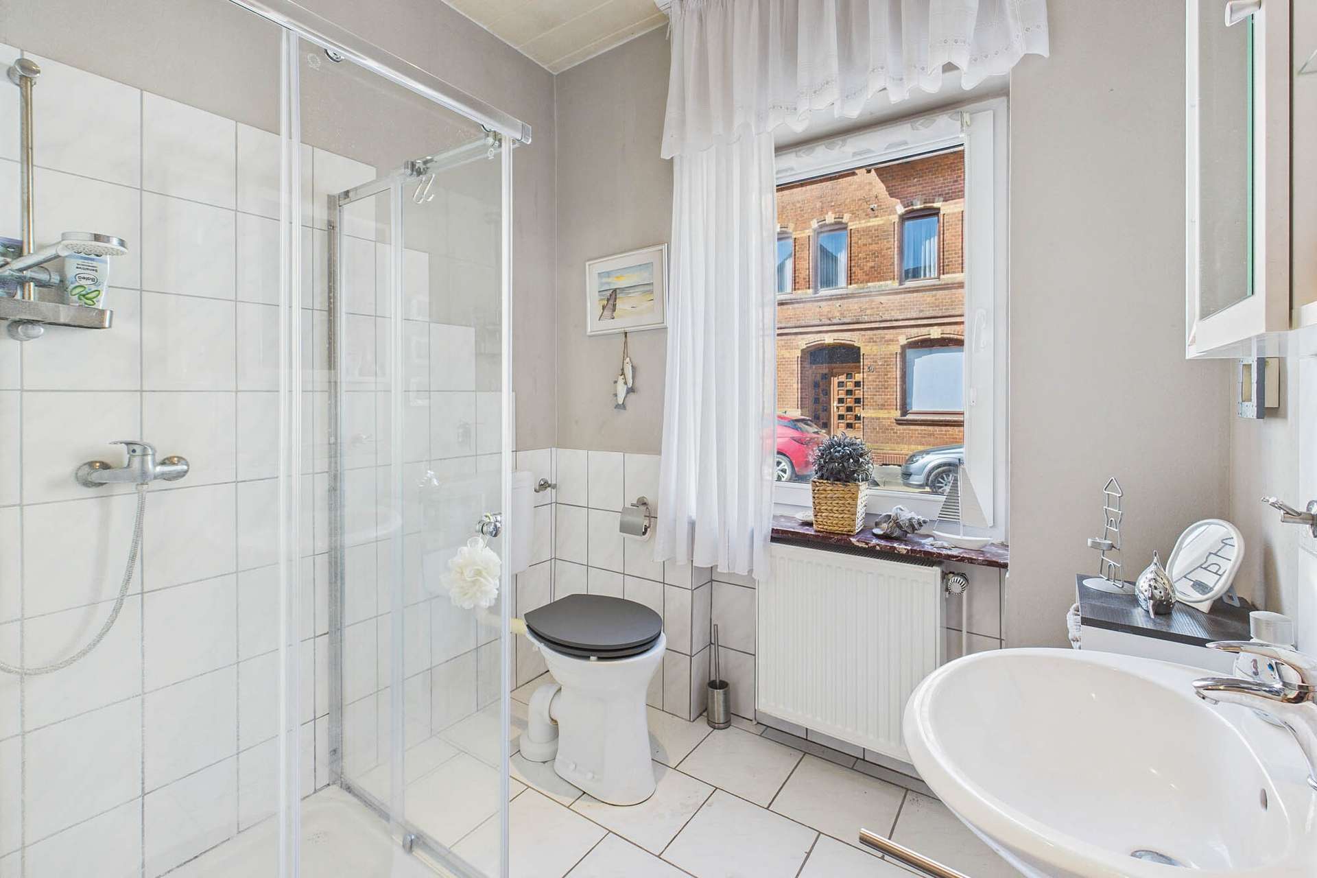 Modernes Badezimmer mit Dusche, WC, Waschbecken und Fensterblick auf eine StraĂe. Helle Fliesen und Fensterdekoration.