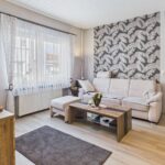 Heller Wohnraum mit beigem Sofa, Holztisch und dekorativer Wand mit Schmetterlingsmuster. Fenster mit VorhÀngen.