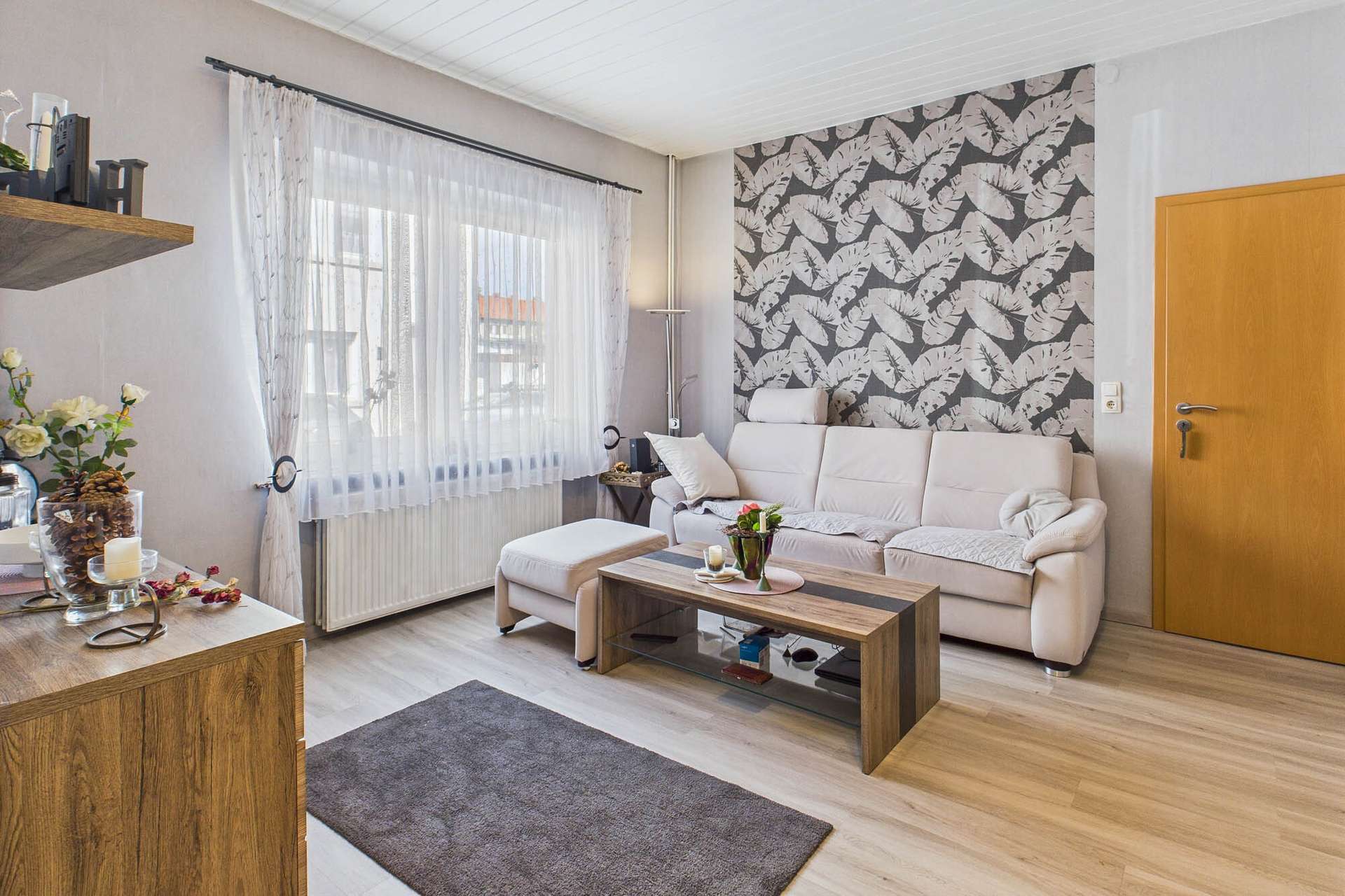 Heller Wohnraum mit beigem Sofa, Holztisch und dekorativer Wand mit Schmetterlingsmuster. Fenster mit VorhÀngen.