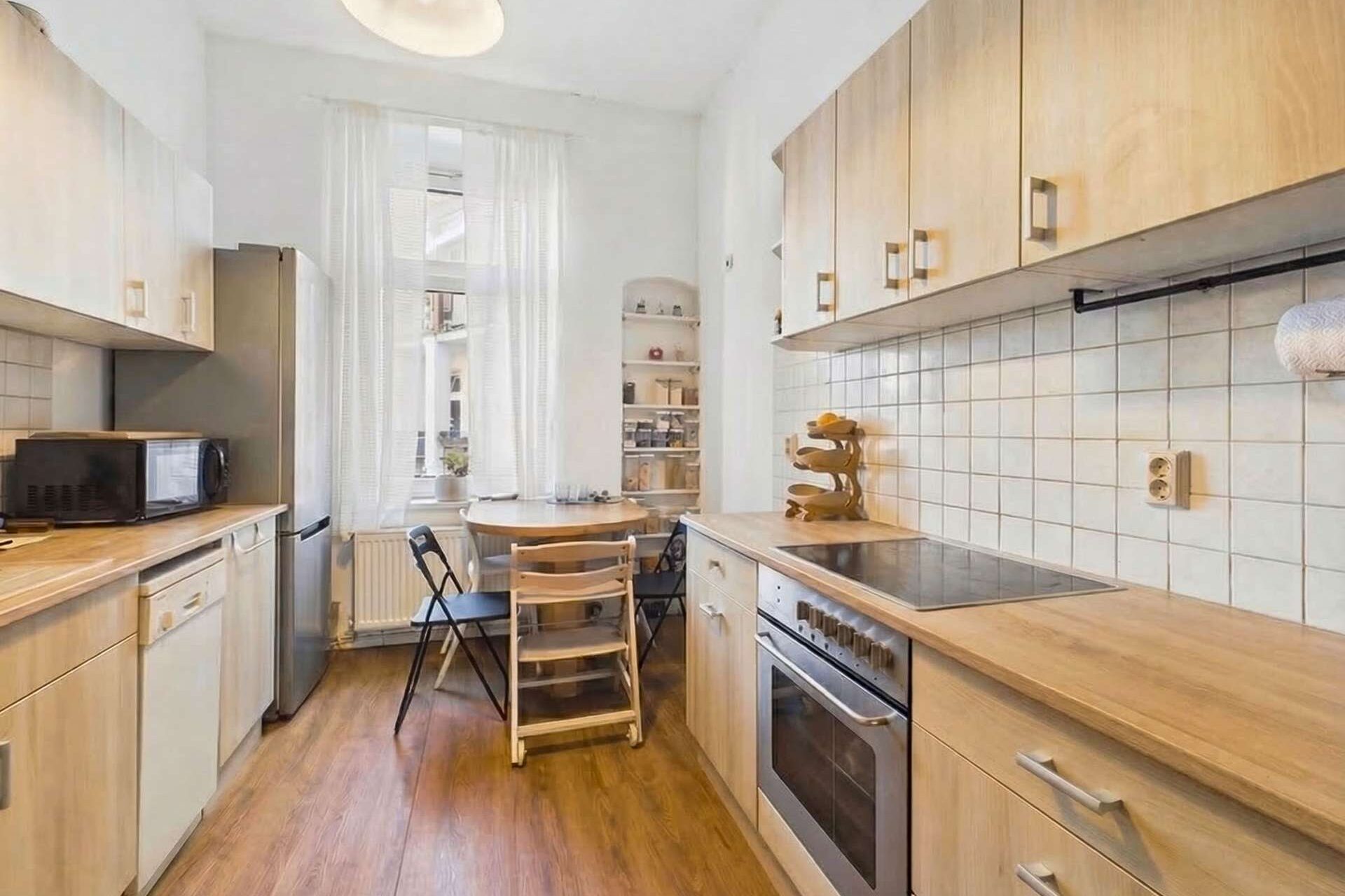 Moderne Küche mit Holzmöbeln, Elektrogeräten, Tisch mit Stühlen und Fenstern, die Tageslicht hereinlassen.